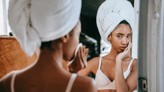 Young woman with towel on head applying skincare in front of a mirror, focusing on her routine.